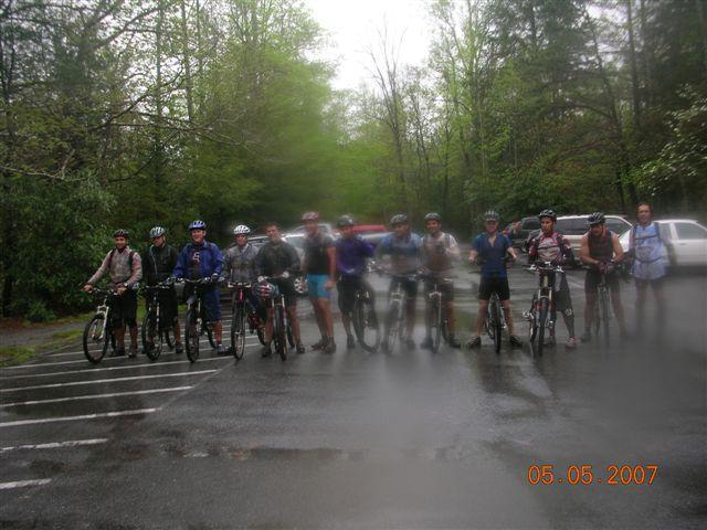A group of bicycle riders, wearing helmets and cycling gear, standing in a row on their mountain bikes in a forested area. The photo appears slightly blurred and misty, likely due to rain, with trees and overcast skies in the background. The date at the bottom reads May 5, 2007. Black Mountain mountain bike trail.