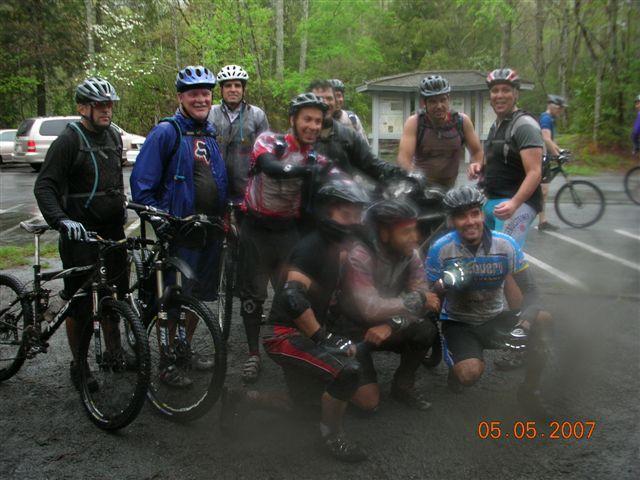 A group of ten mountain bikers posing together in a forested area after a ride. They are wearing helmets and biking gear, some appearing muddy from their adventure. The background features trees and a building, with parked cars visible. The date displayed in the corner is May 5, 2007. Black Mountain mountain bike trail.