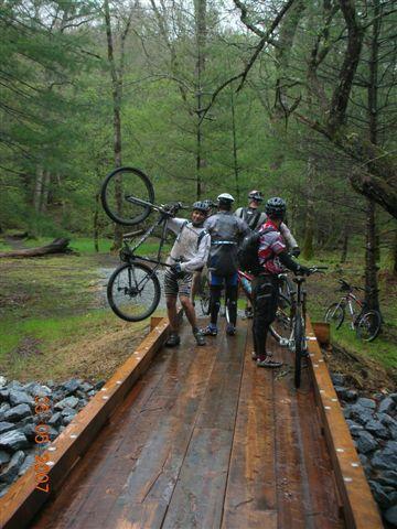 A group of five mountain bikers stands on a wooden bridge in a forested area. One biker is lifting their bike above their head, while the others are gathered around, all wearing helmets and cycling gear. The scene is set in a lush, green environment, indicating a wet and possibly rainy day. Black Mountain mountain bike trail.