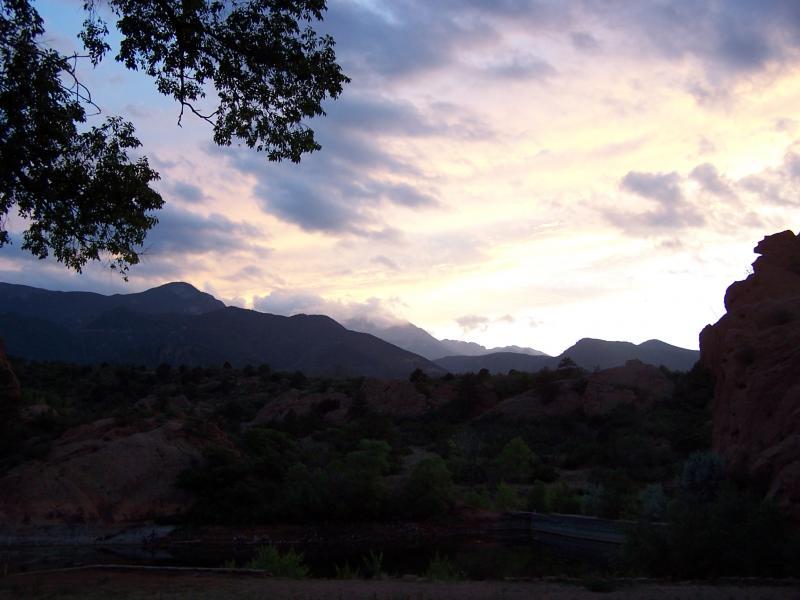 A tranquil landscape at sunset, featuring silhouetted mountains against a colorful sky filled with clouds. In the foreground, trees frame the scene, while a small body of water reflects the evening light, adding to the serene atmosphere. Red Rock Canyon mountain bike trail.