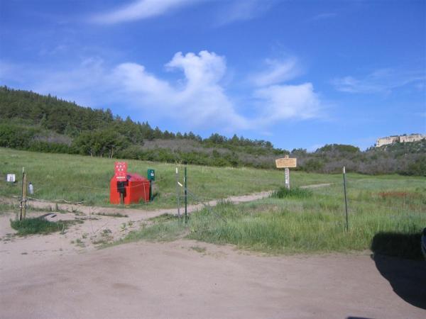 A scenic view of a grassy field under a blue sky with scattered clouds. In the foreground, there is a red information kiosk and a green box, both near a dirt path leading into the field. A wooden sign stands beside the path, indicating directions or information about the area. Lush greenery and trees are visible in the background. Spruce Mountain Trail Upper Loop mountain bike trail.