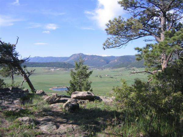 A scenic landscape view showcasing rolling green hills, a small pond, and distant mountains under a clear blue sky, framed by trees in the foreground. Spruce Mountain Trail Upper Loop mountain bike trail.