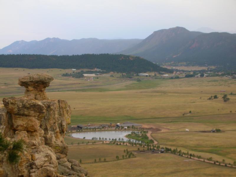 A scenic view of a rocky outcrop in the foreground, with a sprawling landscape of fields and a small pond below. In the background, there are rolling hills and mountains under a gray sky. Spruce Mountain Trail Upper Loop mountain bike trail.