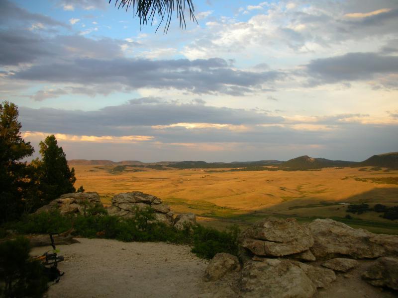 A scenic view of rolling hills under a partly cloudy sky, featuring rocky outcrops in the foreground and expansive grasslands stretching into the distance. The warm light of sunset casts a golden hue over the landscape. Spruce Mountain Trail Upper Loop mountain bike trail.
