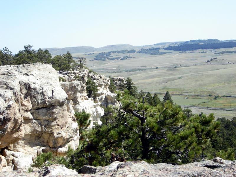 A panoramic view of a rocky landscape with cliffs in the foreground and rolling green hills in the background under a clear blue sky. Pine trees are scattered across the scene, adding greenery to the rocky terrain. Spruce Mountain Trail Upper Loop mountain bike trail.