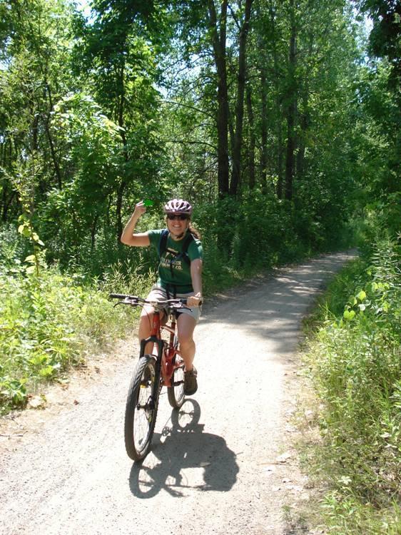 A person riding a mountain bike along a dirt path surrounded by greenery, wearing a helmet and a green shirt, smiling and raising one arm in a gesture of excitement or greeting. Bloomington Ferry Trail mountain bike trail.