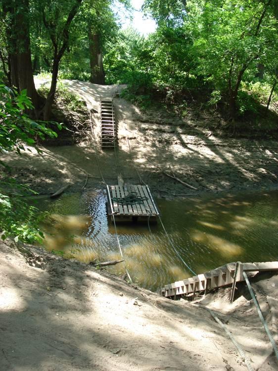 A serene view of a small wooden dock on a shallow body of water, surrounded by lush greenery. A visible sandy path leads up to a set of wooden stairs on the left side, while a rope extends from the dock to the shore. Sunlight filters through the trees, creating a peaceful atmosphere. Bloomington Ferry Trail mountain bike trail.