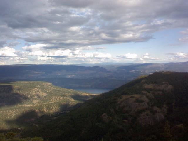 A panoramic view of a mountainous landscape under a partly cloudy sky, showcasing rolling hills, a river, and lush greenery. The scene captures the tranquility of nature from an elevated perspective. Mcdougal Rim mountain bike trail.