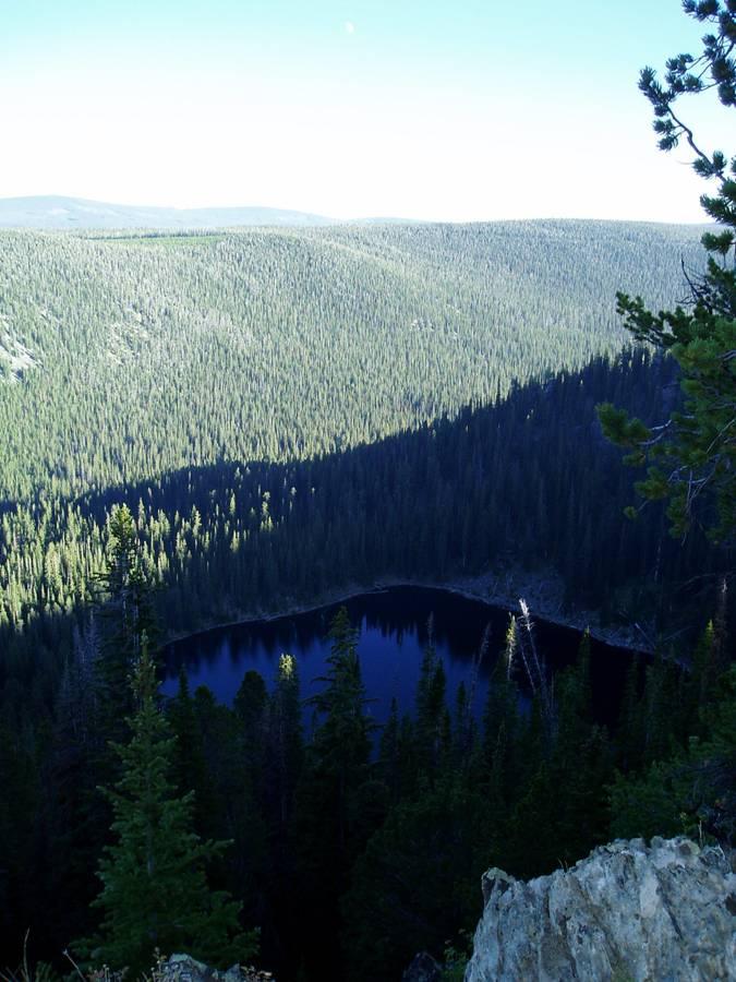 A panoramic view of a forested landscape featuring a dark, reflective body of water nestled among evergreen trees. The scene captures the serene beauty of nature with rolling hills in the background and a clear blue sky above. Sunlight highlights the contours of the landscape, creating a contrast between the shadows and the sunlit areas. Crater Lake mountain bike trail.