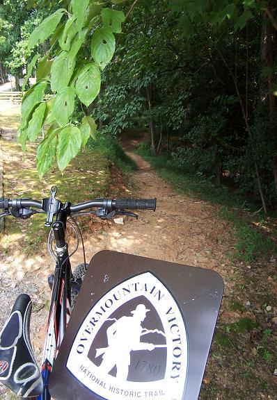 A mountain bike is positioned in the foreground, with its handlebars visible and a sign displayed in front reading "Overmountain Victory National Historic Trail." In the background, a narrow dirt path winds through lush greenery, with trees and foliage framing the scene. Overmountain Victory Trail mountain bike trail.