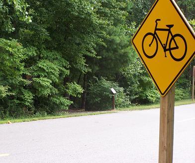 A bicycle warning sign is prominently displayed at the edge of a quiet road lined with dense green foliage, indicating a bike-friendly area or trail nearby. Overmountain Victory Trail mountain bike trail.