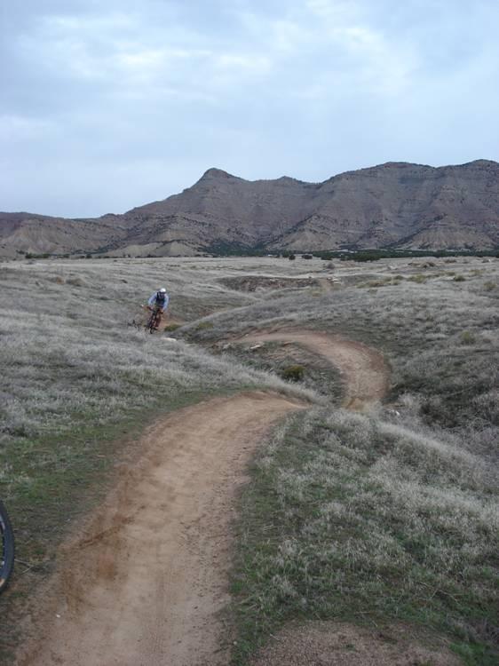 A mountain biker navigating a winding dirt trail in a grassy landscape, with rolling hills and mountains in the background under a cloudy sky. Kessel Run mountain bike trail.