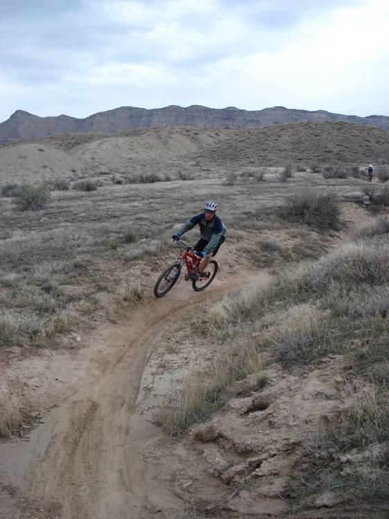 A person riding a mountain bike on a dirt trail in a hilly, outdoor landscape, surrounded by sparse vegetation and rolling hills under a cloudy sky. Kessel Run mountain bike trail.