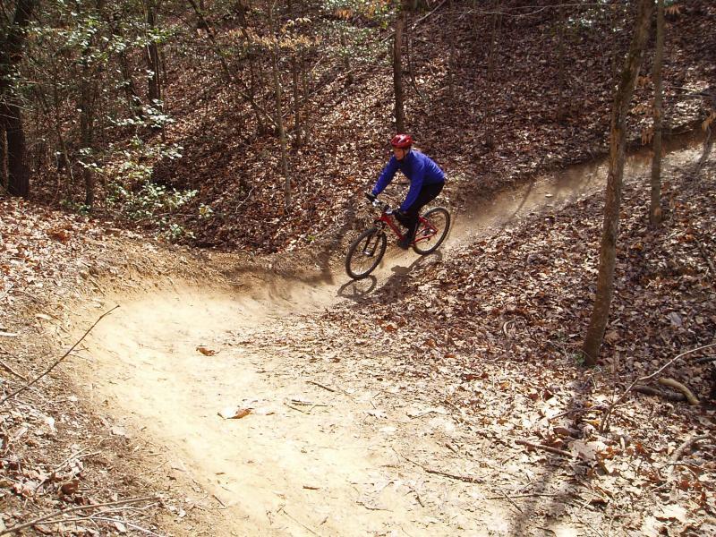 A mountain biker wearing a blue jacket and red helmet rides around a dirt curve on a forest trail, surrounded by trees and fallen leaves. The path shows signs of recent use with a swirl of dust trailing behind the bike. Overmountain Victory Trail mountain bike trail.