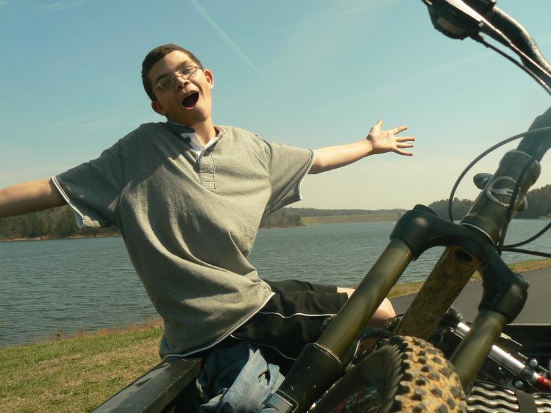 A smiling young person with glasses is seated on a bike, arms spread wide in a joyful pose. The background features a serene lake surrounded by trees under a clear blue sky. The scene conveys a sense of fun and adventure. Overmountain Victory Trail mountain bike trail.