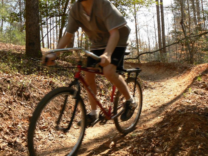 A cyclist riding a red mountain bike on a dirt trail surrounded by trees. The cyclist is wearing a gray shirt and black shorts, captured in motion as they navigate a curved path through a wooded area. Leaves and natural terrain are visible along the trail. Overmountain Victory Trail mountain bike trail.