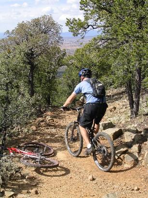 A mountain biker navigating a rocky trail in a forested area, with a second bike laying on the ground nearby. The landscape features trees and distant mountains under a partly cloudy sky. Devisadero Loop (108) mountain bike trail.