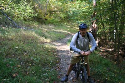 A person in a helmet stands by their mountain bike on a dirt trail surrounded by trees. The scene features autumn foliage, with sunlight filtering through the leaves. The individual is wearing a long-sleeve shirt, shorts, and biking shoes, appearing to take a break from riding. Black Hill Regional Park mountain bike trail.