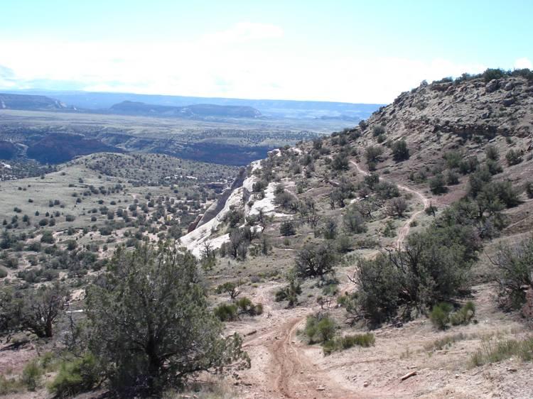 A scenic view of a rugged landscape featuring rolling hills and rocky outcrops under a blue sky with scattered clouds. The foreground includes a dirt path winding through sparse vegetation, while the expansive valley stretches out in the background, dotted with shrubs and trees. Western Rim mountain bike trail.
