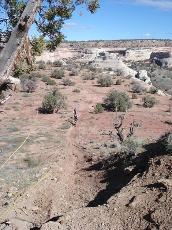 A dusty, rugged landscape with reddish soil and sparse vegetation. In the foreground, a narrow dirt path leads downhill, while a rider on a mountain bike can be seen descending. In the background, dramatic rock formations and cliffs rise against a blue sky dotted with clouds. The scene captures a sense of adventure in a natural outdoor setting. Western Rim mountain bike trail.