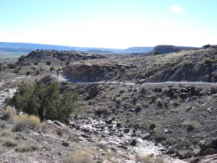 A scenic view of a rocky landscape with rolling hills and sparse vegetation, featuring a person riding a bicycle on a winding dirt path. The sky is clear with a few clouds, and the distant hills create a beautiful backdrop. Western Rim mountain bike trail.