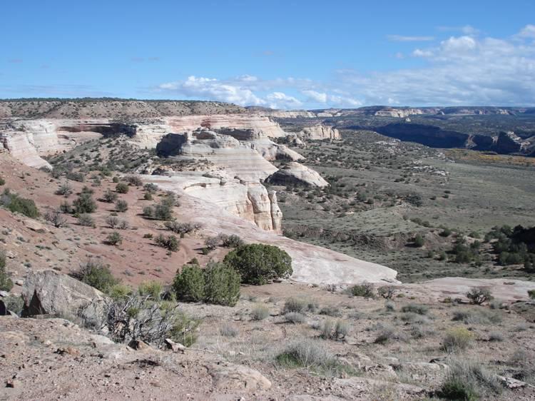 A panoramic view of rugged, multi-layered rock formations and rolling hills under a blue sky, with some scattered vegetation in the foreground. The landscape features a mix of earthy tones and layers, showcasing natural erosion and geological diversity. Western Rim mountain bike trail.