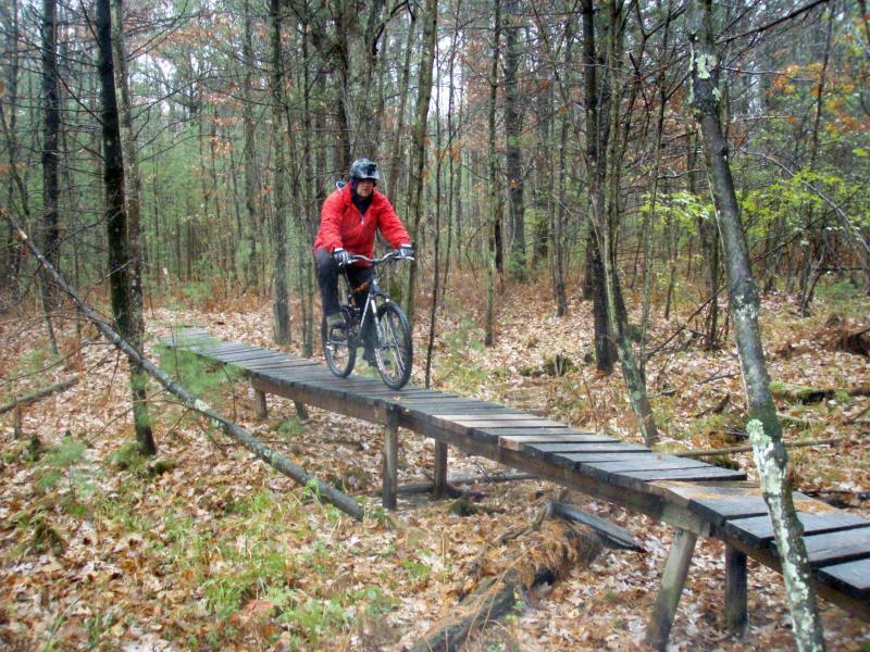 A cyclist in a red jacket rides over a wooden bridge in a forest, surrounded by trees and fallen leaves. The scene appears damp, suggesting recent rain. Levis Mounds mountain bike trail.