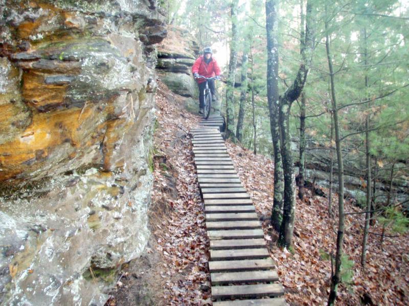 A mountain biker in a red jacket rides along a narrow wooden boardwalk set against a rocky cliff in a forested area. Surrounding trees and fallen leaves create a natural trail environment. Levis Mounds mountain bike trail.