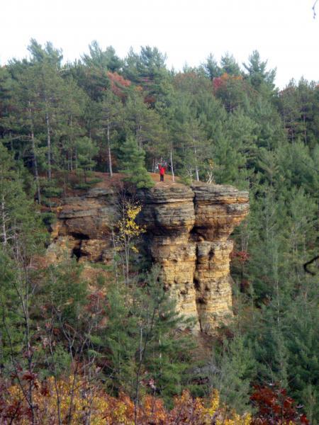 A person standing on top of a large rock formation surrounded by dense pine trees and autumn foliage. Levis Mounds mountain bike trail.