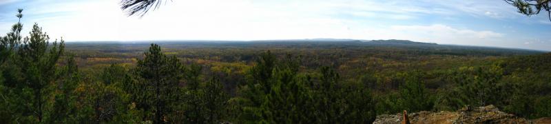 Panoramic view of a lush green landscape with rolling hills and a clear sky, showcasing a dense forest of pines and deciduous trees. The scene captures the vibrant colors of the foliage, indicating a season of change, and extends to the horizon where mountains are faintly visible in the distance. Levis Mounds mountain bike trail.