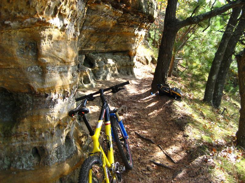 Two mountain bikes, one yellow and one blue, are parked beside a rocky trail surrounded by trees. The path is narrow and lined with natural rock formations and fallen leaves, indicating an outdoor setting suitable for biking and nature exploration. Levis Mounds mountain bike trail.