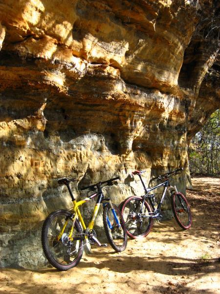 Three mountain bikes in yellow, blue, and black are leaning against a rugged, natural rock formation with warm tones and textures. The sandy ground and surrounding trees provide a serene outdoor setting for outdoor activities. Levis Mounds mountain bike trail.