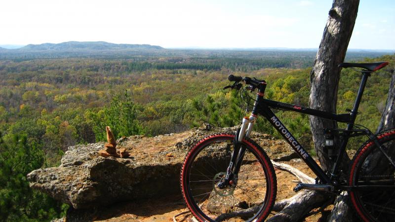 A mountain bike resting on a rocky outcrop, overlooking a vast landscape of trees and rolling hills under a bright blue sky. The scene captures the beauty of nature and an ideal spot for outdoor adventure. Levis Mounds mountain bike trail.