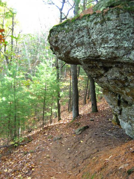 A rocky outcrop extends over a footpath in a forested area, surrounded by tall trees and patches of green foliage. Fallen leaves cover the ground, indicating an autumn setting. The sunlight filters through the trees, creating a serene natural atmosphere. Levis Mounds mountain bike trail.