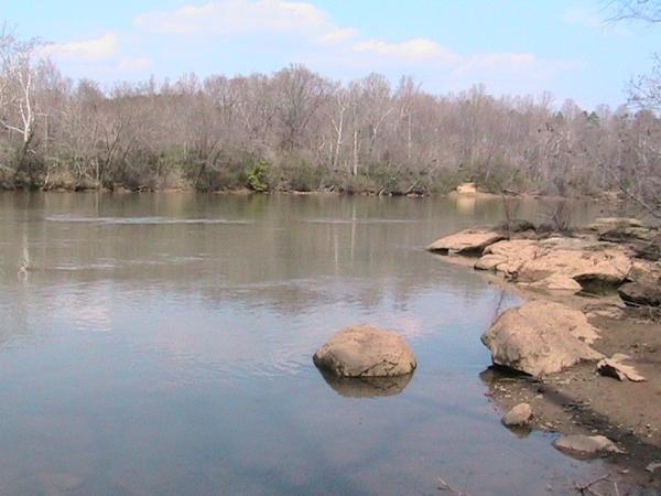 A peaceful river scene featuring calm water reflecting the sky, with rocky shores and bare trees lining the banks. The landscape is serene, capturing the tranquility of nature in a rural setting. Riverbend Park mountain bike trail.
