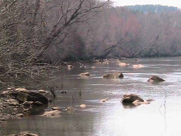 A calm river scene surrounded by bare trees, with large rocks partially submerged in the water. The landscape is serene, depicting a tranquil natural setting under overcast skies. Riverbend Park mountain bike trail.