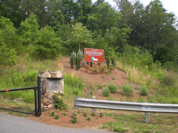 Sign at the entrance of River Bend Park, surrounded by greenery and plants, with a stone gate column and a metal fence in the foreground. Riverbend Park mountain bike trail.