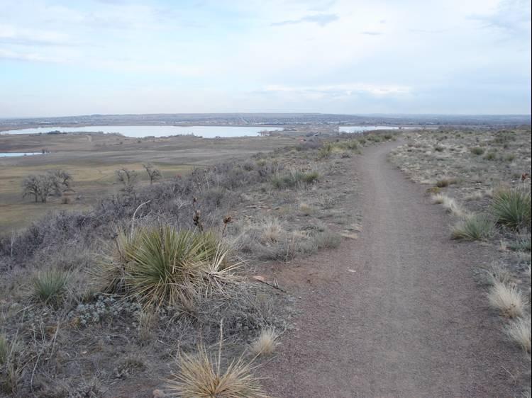 A winding dirt path leads through a grassy landscape, with sparse vegetation and shrubs. In the distance, a body of water can be seen, surrounded by open fields under a cloudy sky. The scene captures a serene and tranquil outdoor setting. Boulder Valley Ranch mountain bike trail.