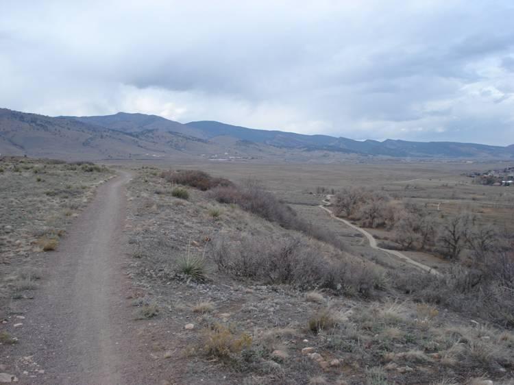 A winding dirt trail leads through a sparse, grassy landscape with rolling hills and distant mountains under a cloudy sky. The scene conveys a sense of tranquility and openness, with a few scattered trees visible along a nearby path. Boulder Valley Ranch mountain bike trail.
