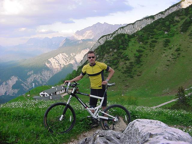 A cyclist wearing a yellow and black jersey stands next to a mountain bike on a grassy hillside, with rocky terrain and mountains in the background. A building is visible in the distance, under a partly cloudy sky. Garmisch To Osterfelder Bergstation mountain bike trail.