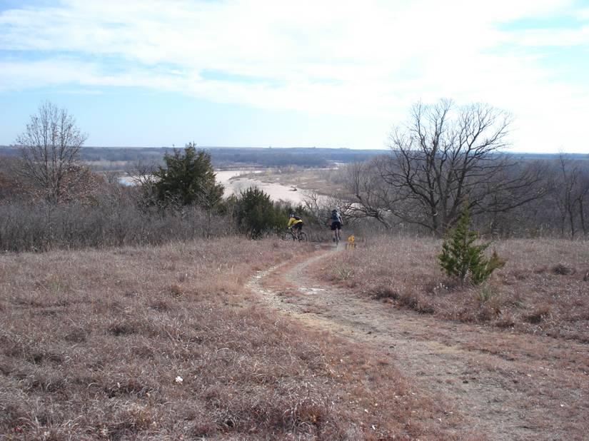 Two cyclists ride along a winding dirt path on a hillside, surrounded by dry grass and sparse trees. In the background, a riverbed is visible, stretching across a landscape under a partly cloudy sky. The scene captures a peaceful outdoor setting, ideal for hiking or biking. Camp Horizon mountain bike trail.