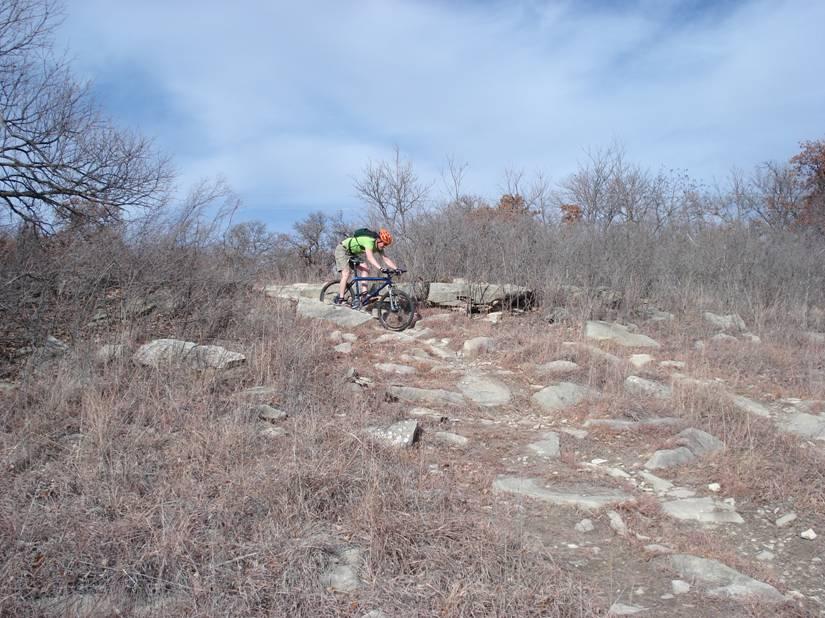 A mountain biker navigating rocky terrain on a trail surrounded by sparse vegetation and trees under a cloudy sky. Camp Horizon mountain bike trail.