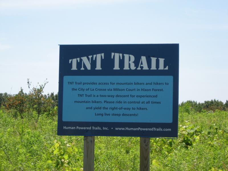 Sign for the TNT Trail located in Hixon Forest, providing access for mountain bikers and hikers. The sign includes information about the trail's two-way descent, guidelines for riders to maintain control, and a reminder to yield the right-of-way to hikers. The background features greenery typical of a forested area. Hixon Forest mountain bike trail.