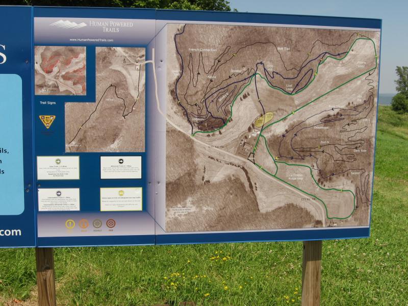 A trail map displayed on a large sign, featuring various trails marked in green and blue, alongside a legend with symbols. The map outlines the terrain and pathways for human-powered outdoor activities, with a grassy area in the foreground and trees in the background. Hixon Forest mountain bike trail.