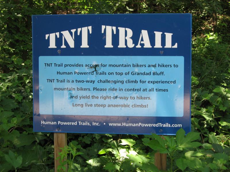 Blue sign for the TNT Trail, providing information for mountain bikers and hikers. The sign states that the trail offers access on top of Grandad Bluff, describes it as a two-way challenging climb suited for experienced bikers, and emphasizes the importance of yielding to hikers while riding in control. The bottom of the sign includes the name and website of Human Powered Trails, Inc. The background features lush greenery. Hixon Forest mountain bike trail.