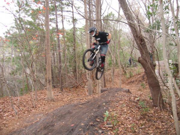 A mountain biker in a helmet performs a jump off a dirt mound in a wooded area, surrounded by trees and autumn foliage. Blue Clay Bike Park mountain bike trail.