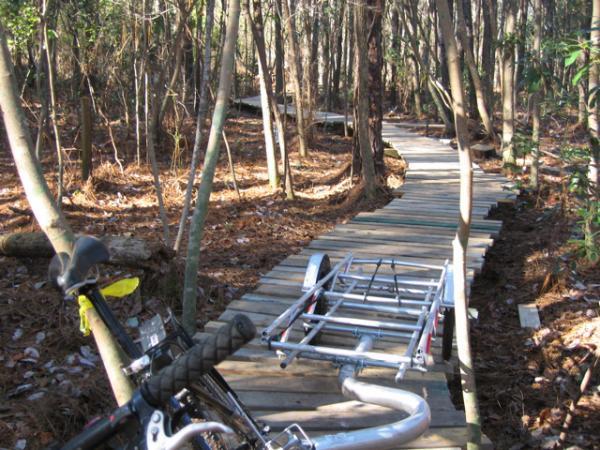 A narrow wooden boardwalk winding through a forest path, surrounded by trees and fallen leaves. A bicycle is leaning against the side of the boardwalk, with a trailer attached nearby. The scene captures a quiet, natural setting ideal for outdoor activities. Blue Clay Bike Park mountain bike trail.