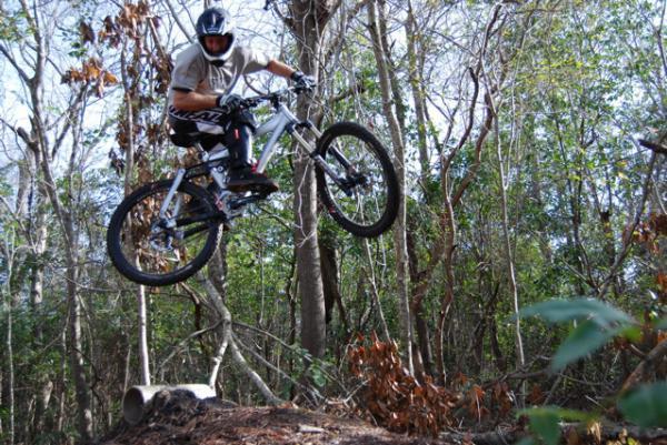 A mountain biker performing a jump over a log, surrounded by trees and greenery in a forested area. The cyclist is in mid-air, wearing a helmet and protective gear, showcasing dynamic movement and skill. Blue Clay Bike Park mountain bike trail.