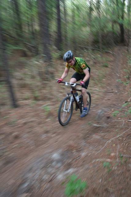 A mountain biker in motion navigating a dirt trail through a forested area, wearing a helmet and a colorful jersey. The background shows blurred trees and ground, indicating speed and action. Blue Clay Bike Park mountain bike trail.