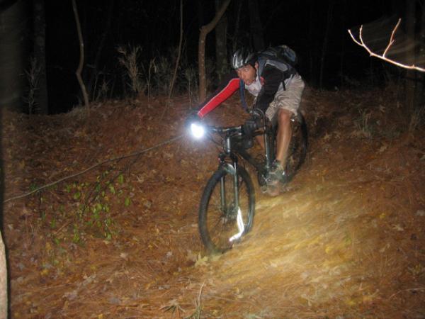 A mountain biker navigating a forest trail at night, wearing a helmet and a backpack, with a bright front light illuminating the path. The surroundings are filled with pine needles and trees, creating a rugged and adventurous atmosphere. Blue Clay Bike Park mountain bike trail.
